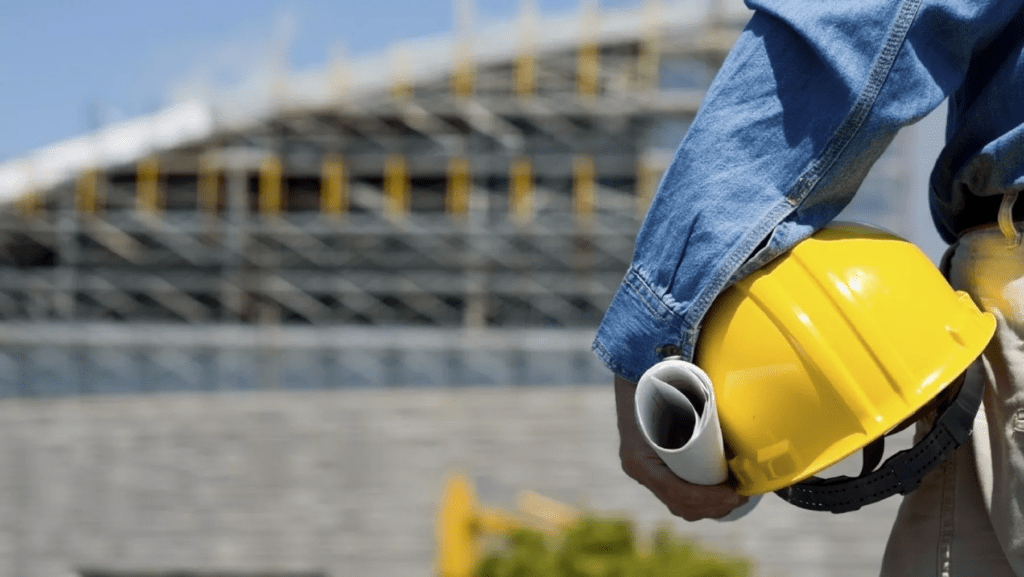 Un lavoratore con casco protettivo e il disegno di un progetto edilizio in mano; si avvia verso un edificio in costruzione.