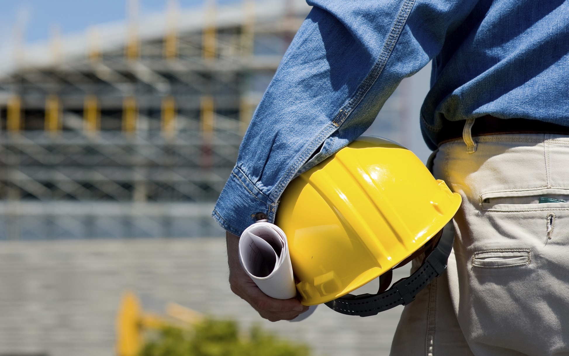 Uomo di spalle che guarda di fronte a sé un cantiere e regge in mano un casco di protezione e carte.