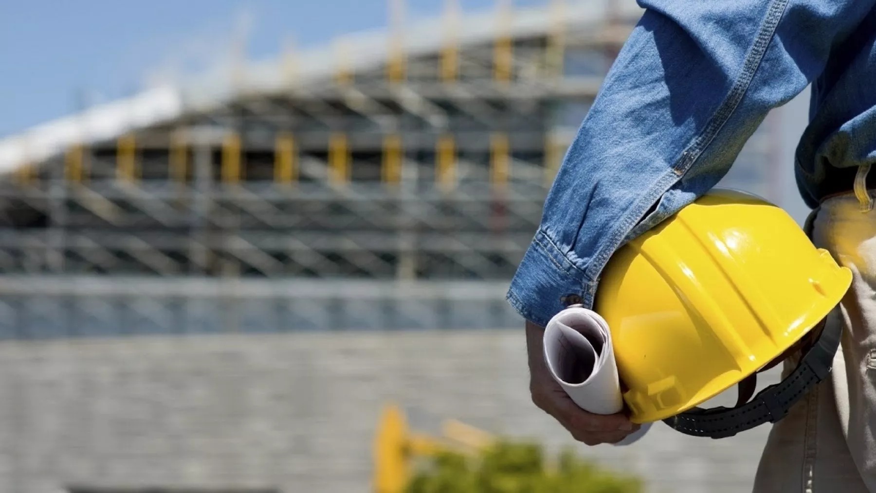 Un lavoratore con casco protettivo e un progetto edilizio in mano; si avvia verso un edificio in costruzione.
